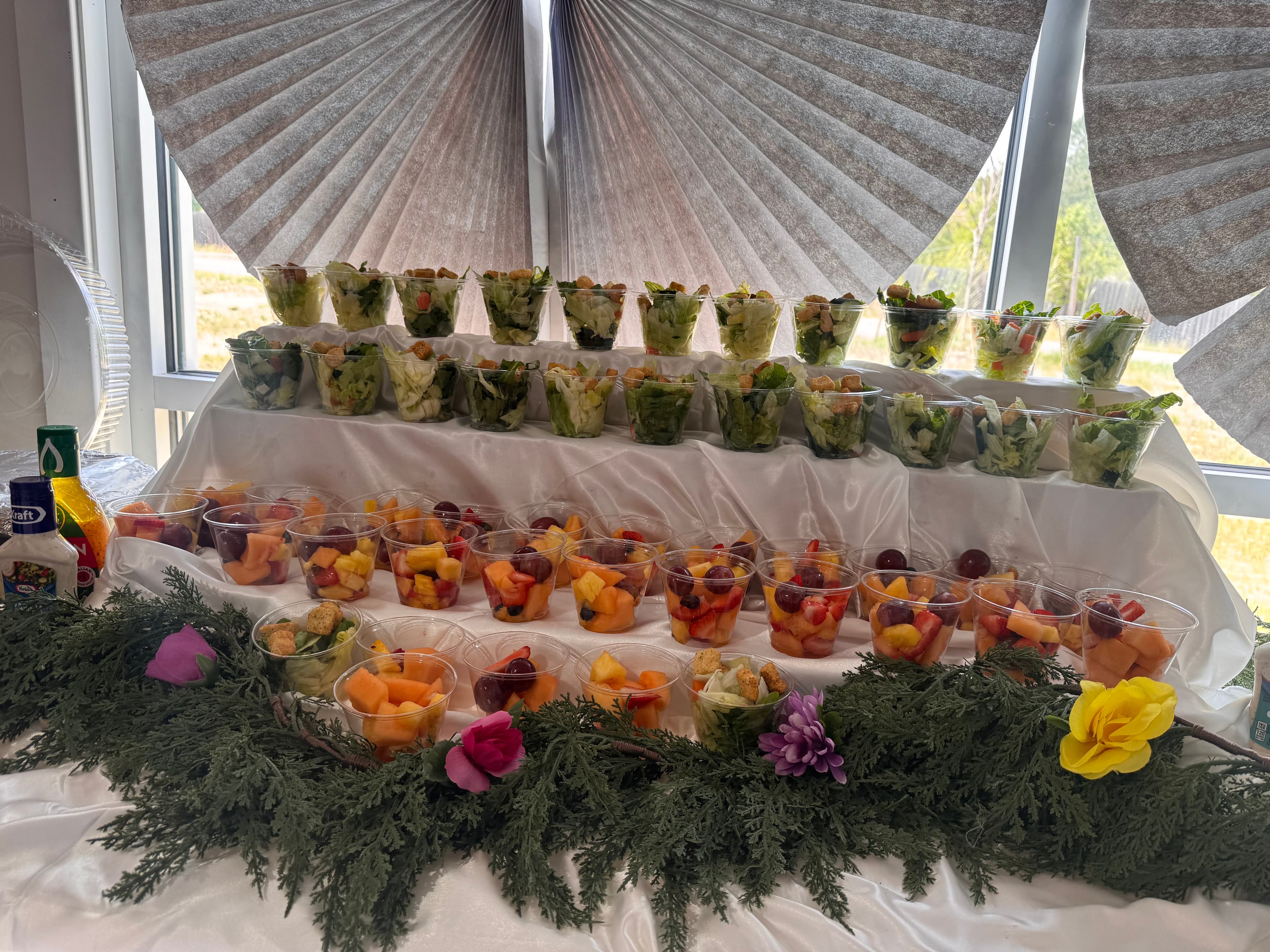 Individual salad and fruit cups on a tiered display decorated with greenery and flowers.